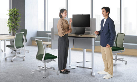 新商品アセンドの紹介画像。台形天板の昇降テーブル。高さを上げて、立ってパソコンワークをしている女性と、横に立って話しかけている男性。